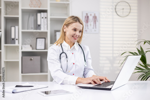 Mature female doctor working inside medical office, pediatrician using laptop while sitting inside clinic office, female worker in medical gown smiling.