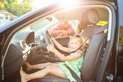 Little girl sitting in driver's seat of car wearing seatbelt, pretending to be old enough to drive and showing she knows the importance of a seat belt as her smiling father watches on on a sunny summe photo