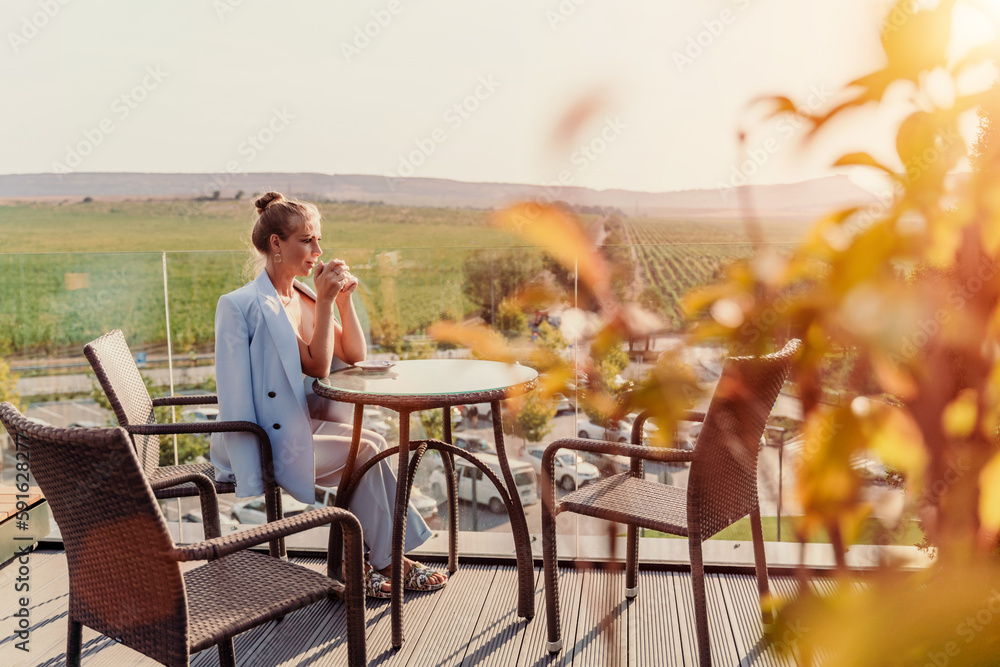 A middle-aged woman sits in a street cafe overlooking the mountains at sunset. She is dressed in a blue jacket and drinks coffee admiring the nature. Travel and vacation concept. A middle-aged woman sits in a street cafe overlooking the mountains at sunset. She is dressed in a blue jacket and drinks coffee admiring the nature. Travel and vacation concept.