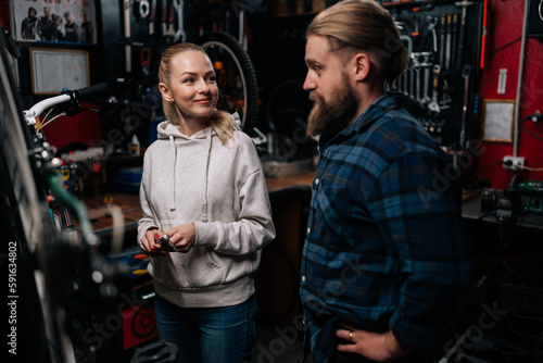 Bearded repairman male communicating with smiling blonde female client, talking problem of bicycle, detected during diagnostics in repair shop with dark interior, standing by MTB.