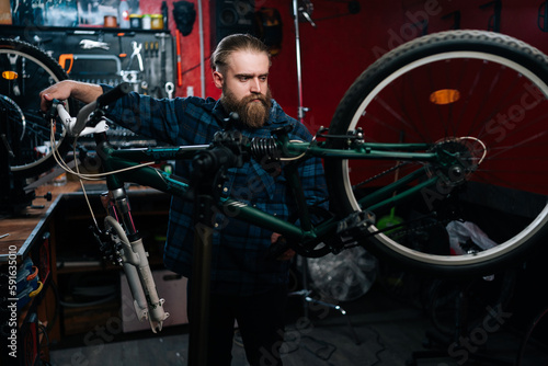 Concentrated bearded repairman watching rear shifter of mountain bike, changing speeds using handlebar shift lever working in bicycle repair shop with dark interior. Concept of maintenance of bicycle.