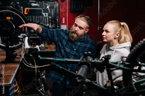 Bearded cycling repairman communicating with blonde female client, talking about problem of bicycle detected during diagnostics in repair shop with dark interior. Concept of bike maintenance.