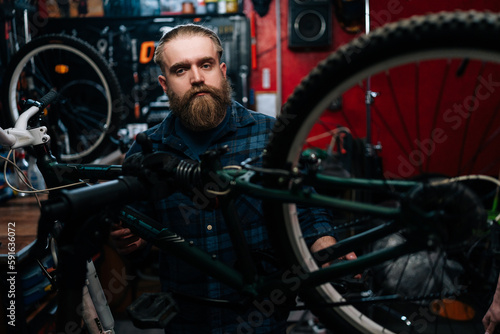 Portrait of handsome bearded cycling repairman with fashion haircut standing by bicycle in repair workshop with dark interior, looking at camera. Concept of professional maintenance of bicycle. © dikushin