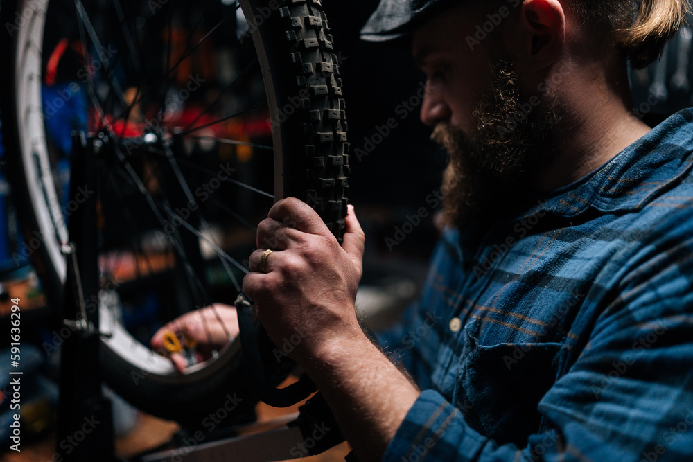 Closeup of bearded repairman using tool to tension spokes in rim of bicycle wheel working in bike repair shop with dark interior. Concept of professional repair and maintenance of bicycle transport.