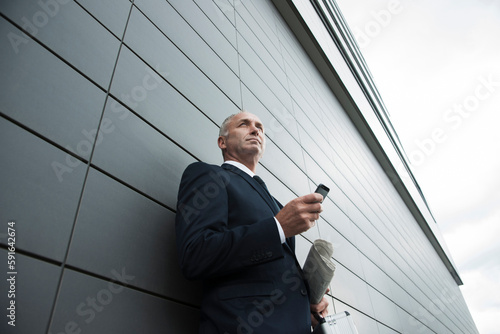 Portrait of businessman leaning against wall of building, holding cell phone, Mannheim, Germany photo