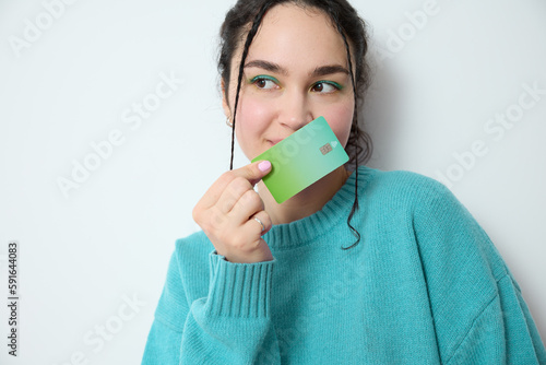 Excited gen z woman with vivid make-up green eye shadow holds a green credit card standing on white backdrop in studio smiles as she showcases her preference of electronic banking and online shopping.