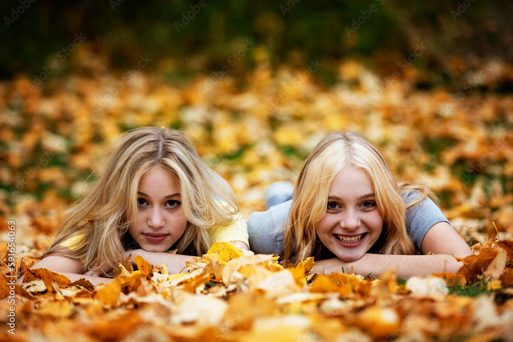 Portrait of two teenage girls laying in the leaves and posing for the camera in a city park on a warm fall afternoon; St. Albert, Alberta, Canada. Portrait of two teenage girls laying in the leaves and posing for the camera in a city park on a warm fall afternoon; St. Albert, Alberta, Canada.