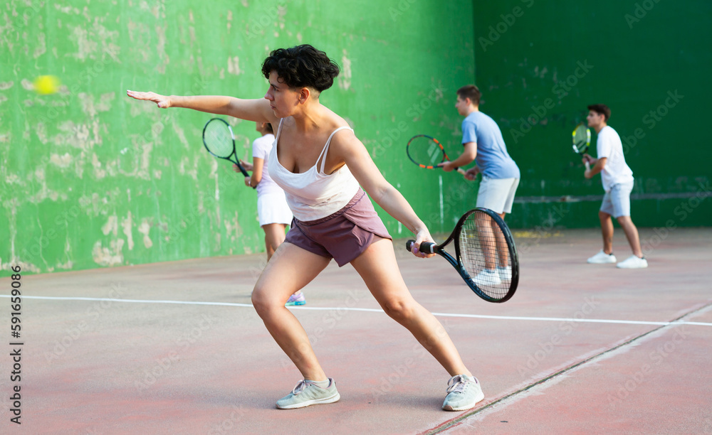 Hispanic woman pelota player hitting ball with racket during training game on outdoor Basque pelota fronton.