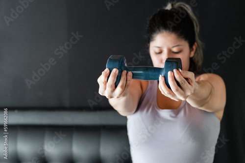 young latina woman very concentrated lifting a blue dumbbell, working her muscle endurance.