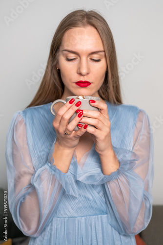 Beautiful young serious woman in light blue dress  with gorgeous long hair  red lips and nails  ring finger  looking at coffee with whipped cream cap.