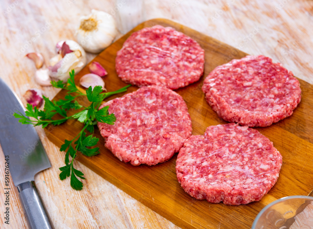 Raw formed hamburger patties ready for cooking on wooden cutting board with fresh fragrant parsley, spicy garlic and allspice