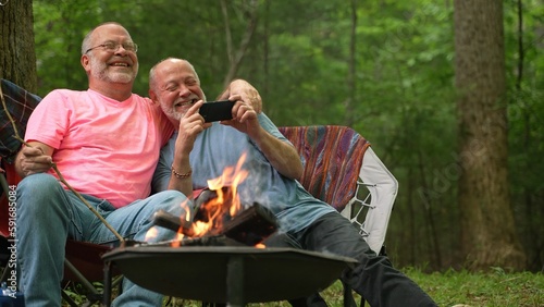 Closeup of two gay men with pride flag and tent in front of campfire talking to smartphone on video chat or taking selfie and kiss. © Robert Peak