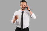 Handsome businessman with cup of coffee talking by mobile phone on grey background
