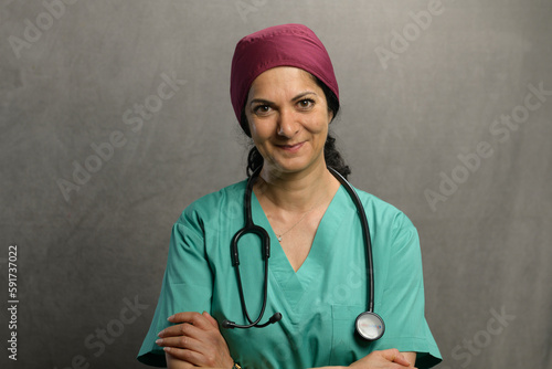 Health professional poses in studio wearing green scrubs, a purple head cover and equipped with a stethoscope.
