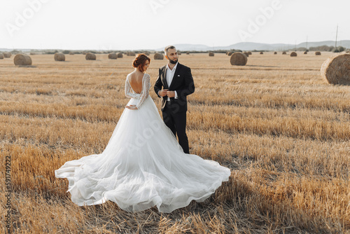 The bride and groom are standing in the field, and behind them are large sheaves of hay. The bride stands with her shoulders turned to the camera. Long elegant dress. Stylish groom. Summer © Vasil
