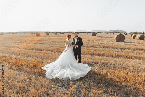 The bride and groom are standing in the field, and behind them are large sheaves of hay. The bride stands with her shoulders turned to the camera. Long elegant dress. Stylish groom. Summer © Vasil