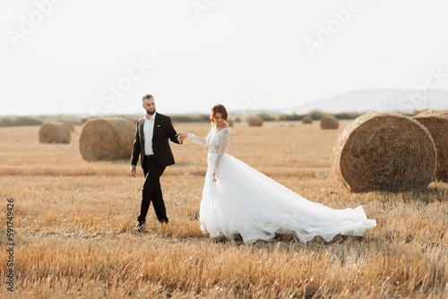 Wedding portrait of the bride and groom walking on the background of hay. The groom holds the hand of the bride and looks at her, her gaze down. Red-haired bride in a long dress. Stylish groom. Summer