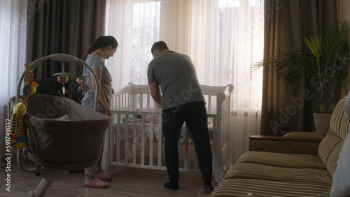 Caucasian couple prepare child crib for little newborn at cozy home. Happy parents put beautiful daughter or son in baby bed, cover with blanket. Concept of childhood, parenthood, love and family.