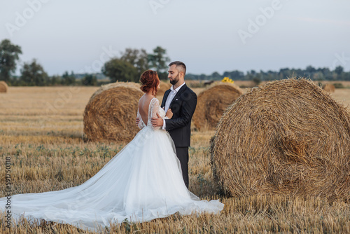 The bride and groom are standing in the field, and behind them are large sheaves of hay. The bride stands with her shoulders turned to the camera. Long elegant dress. Stylish groom. Summer
