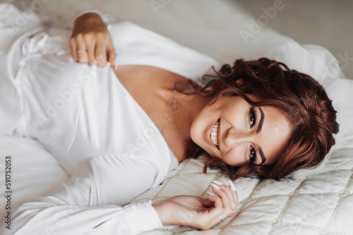 Beautiful young bride in white peignoir lying on bed in hotel bedroom. Final preparations for the wedding. The bride is waiting for the groom. Morning, bride.