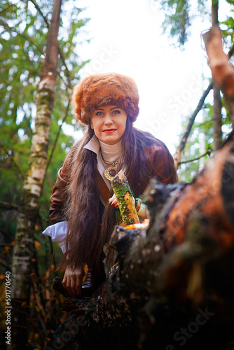 Girl in a leather jacket, a big red fox fur hat in the forest in autumn. A female model poses as a fabulous royal huntress on nature hunt at photo shoot