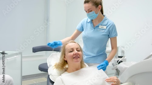 nurse in dental clinic puts on woman bib napkin for patient in dental white background of latest technology excellent light doctor nurse patient preparing for procedure pull up glass table with tools photo