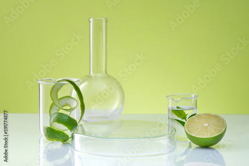 Halves of lime and spiral peel on green background with lab glassware. Petri dish upside down form an empty platform for display cosmetic product of lime extract. Concept laboratory and research.