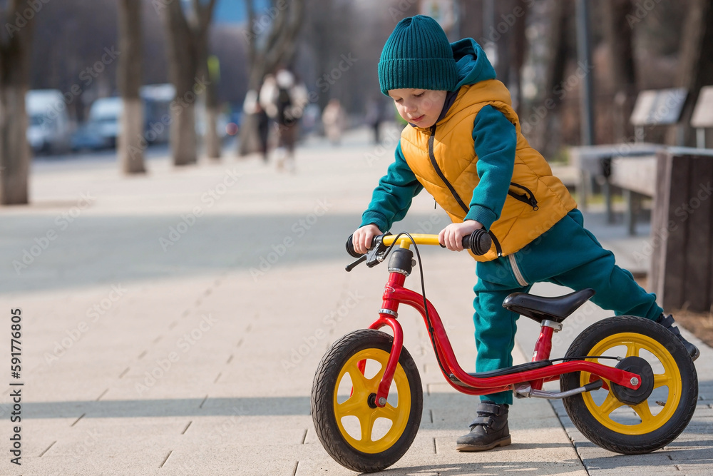 A cheerful little boy rides a bicycle outdoors. A happy child walks in the spring park. The baby is dressed in a fashionable yellow vest and turquoise jumpsuit. - obrazy, fototapety, plakaty 