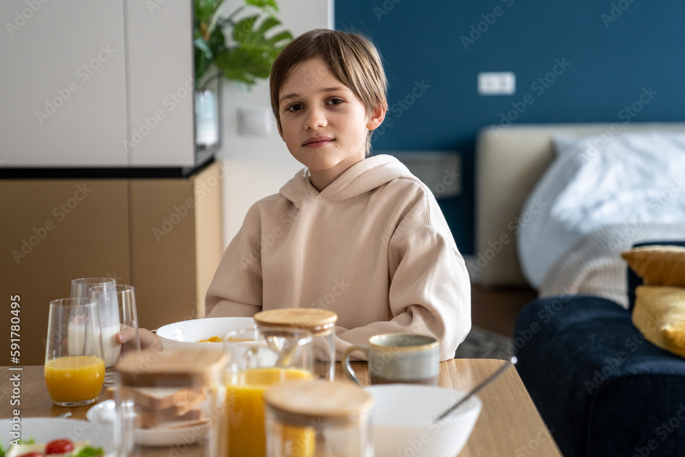 Portrait of happy small european boy having breakfast at home. Smiling little child schoolboy sitting at kitchen table in morning. Interested kid boy looking at camera eating cornflakes before school Portrait of happy small european boy having breakfast at home. Smiling little child schoolboy sitting at kitchen table in morning. Interested kid boy looking at camera eating cornflakes before school