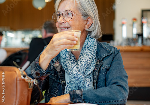 Portrait Senior white haired woman sitting at cafe table holding a coffee and milk glass, elderly lady in denim jacket smiling looking at her side