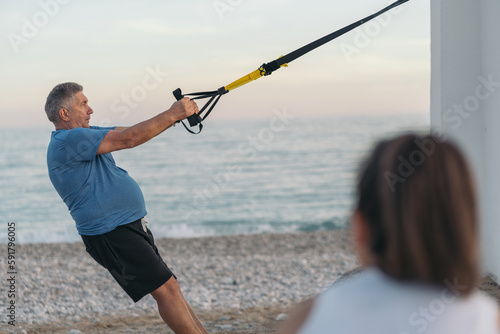 White-haired, overweight older gentleman performing back push-up exercises with suspension training on the beach under the watchful eye of his young personal trainer.