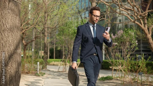 Man in formal suit using phone while Walking at city park. Serious businessman texting on cellphone on the way to office