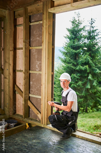 Carpenter constructing wooden framed house. Man worker working with screwdriver, wearing work overalls and helmet. Concept of modern eco-friendly construction.