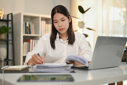 Attractive female accountant holding pen writing on documents, checking marketing report at desk