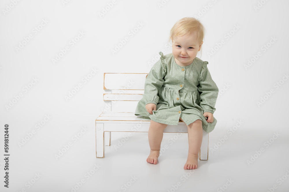Portrait of smiling little barefoot girl with blue eyes wearing green cotton dress sitting on white wooden little bench. Portrait of smiling little barefoot girl with blue eyes wearing green cotton dress sitting on white wooden little bench.