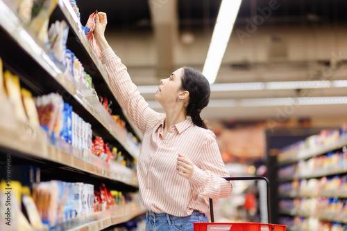 Young caucasian housewife takes food from upper shelve. Side view. Showcase at background. Concept of shopping in grocery store and consumerism photo