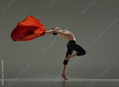 Young flexible, handsome man, shirtless ballet dancer making performance with red silk fabric on grey studio background. Concept of art, classical dance, inspiration, creativity, beauty, choreography