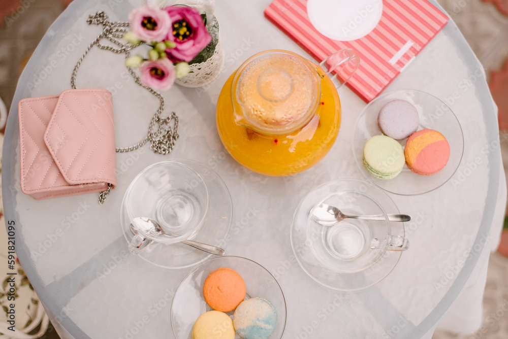 Summer cafe table. On the table are glass plates, cups, a kettle with juice, tea utensils. There are macaroons of different colors in the plates..On the table is a woman's handbag and a notebook Summer cafe table. On the table are glass plates, cups, a kettle with juice, tea utensils. There are macaroons of different colors in the plates..On the table is a woman's handbag and a notebook