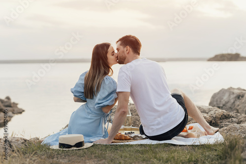 Happy young couple having romantic picnic near the lake. They kissing and clinking wine glasses.