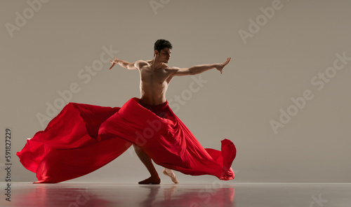 Young, talented, handsome man, ballet dancer dancing with red silk fabric against grey studio background. Passion. Art, classical dance, inspiration, creativity, fashion, beauty, choreography concept