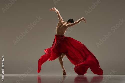 Impressive, artistic young man, professional ballet dancer dancing with red silk fabric against grey studio background. Concept of art, classical dance, inspiration, creativity, beauty, choreography