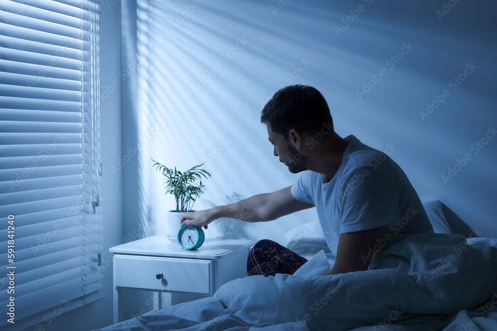 Sleepy man turning off alarm clock on nightstand in morning