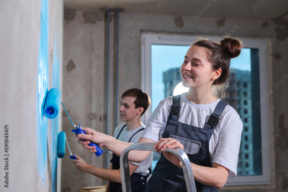 Couple in new home during repair works painting wall together. Happy family holding paint roller painting wall with blue color paint in new house. Home renovation DIY renew home concept Couple in new home during repair works painting wall together. Happy family holding paint roller painting wall with blue color paint in new house. Home renovation DIY renew home concept