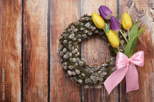 Wreath made of beautiful willow, colorful tulip flowers and pink bow on wooden background, top view. Space for text