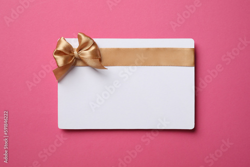 Blank gift card with golden bow on pink background, top view. Space for text