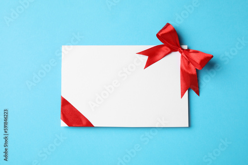 Blank gift card with red bow on light blue background, top view. Space for text
