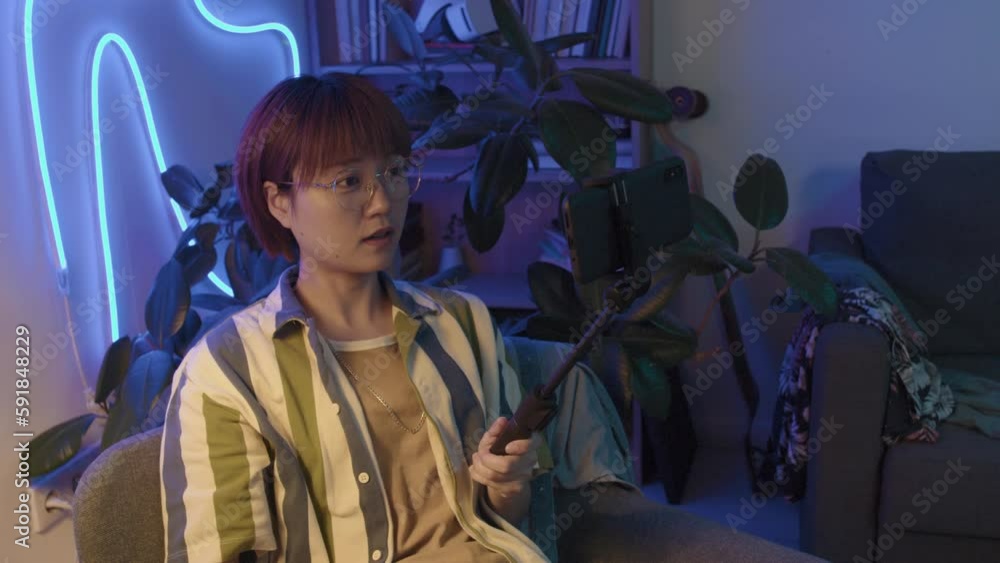 Waist-up shot of young Asian female influencer in casual clothes and glasses recording video content for social media on smartphone with selfie stick, with blue neon lighting in background Waist-up shot of young Asian female influencer in casual clothes and glasses recording video content for social media on smartphone with selfie stick, with blue neon lighting in background