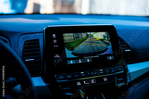 Interior of modern minivan with blurry steering wheel, large display show AC temperature, backup camera guide lines, wooden fence along residential street back alley