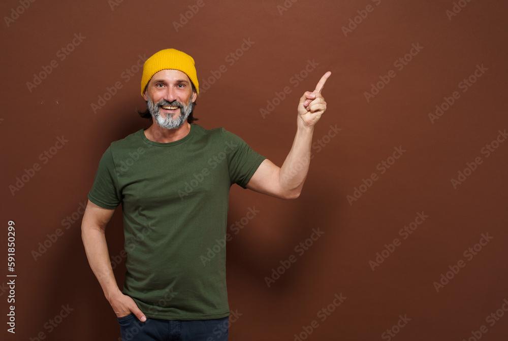 Smiling European man in a yellow hat points his hand to the side with copy space on a brown background. Product placing, advertising concept.