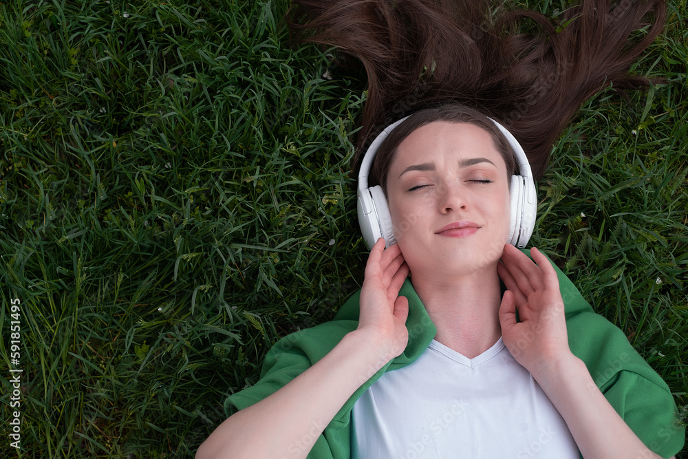 A teenage girl lies on the grass in white headphones, her eyes are closed, listening to music, taking a break from the hustle and bustle, dressed in a white T-shirt, a green jacket is thrown over A teenage girl lies on the grass in white headphones, her eyes are closed, listening to music, taking a break from the hustle and bustle, dressed in a white T-shirt, a green jacket is thrown over